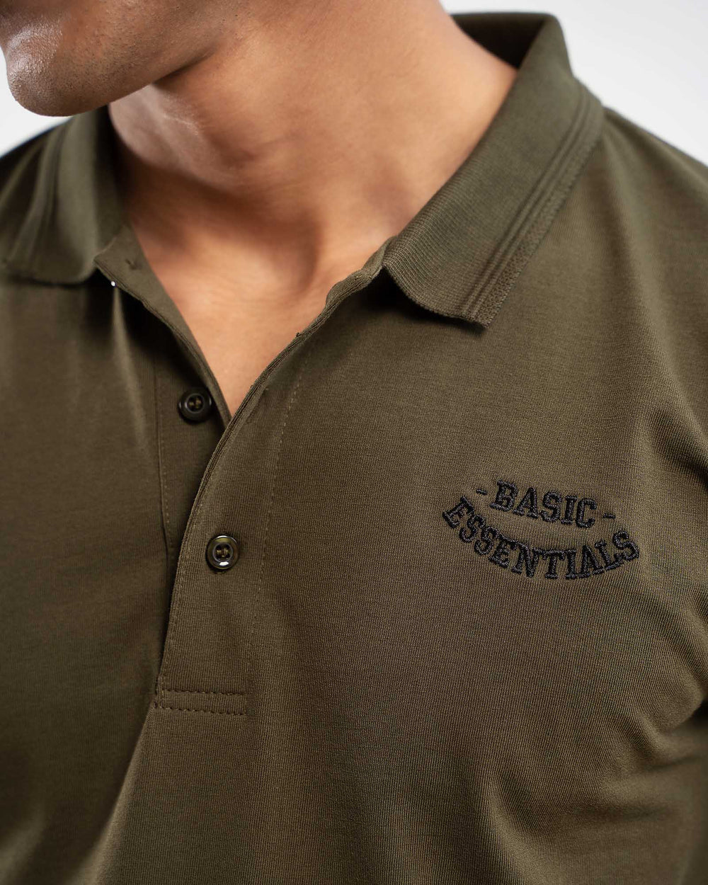 Close-up of olive green polo shirt collar with small dark embroidered "Basic Essentials" arched text logo on chest