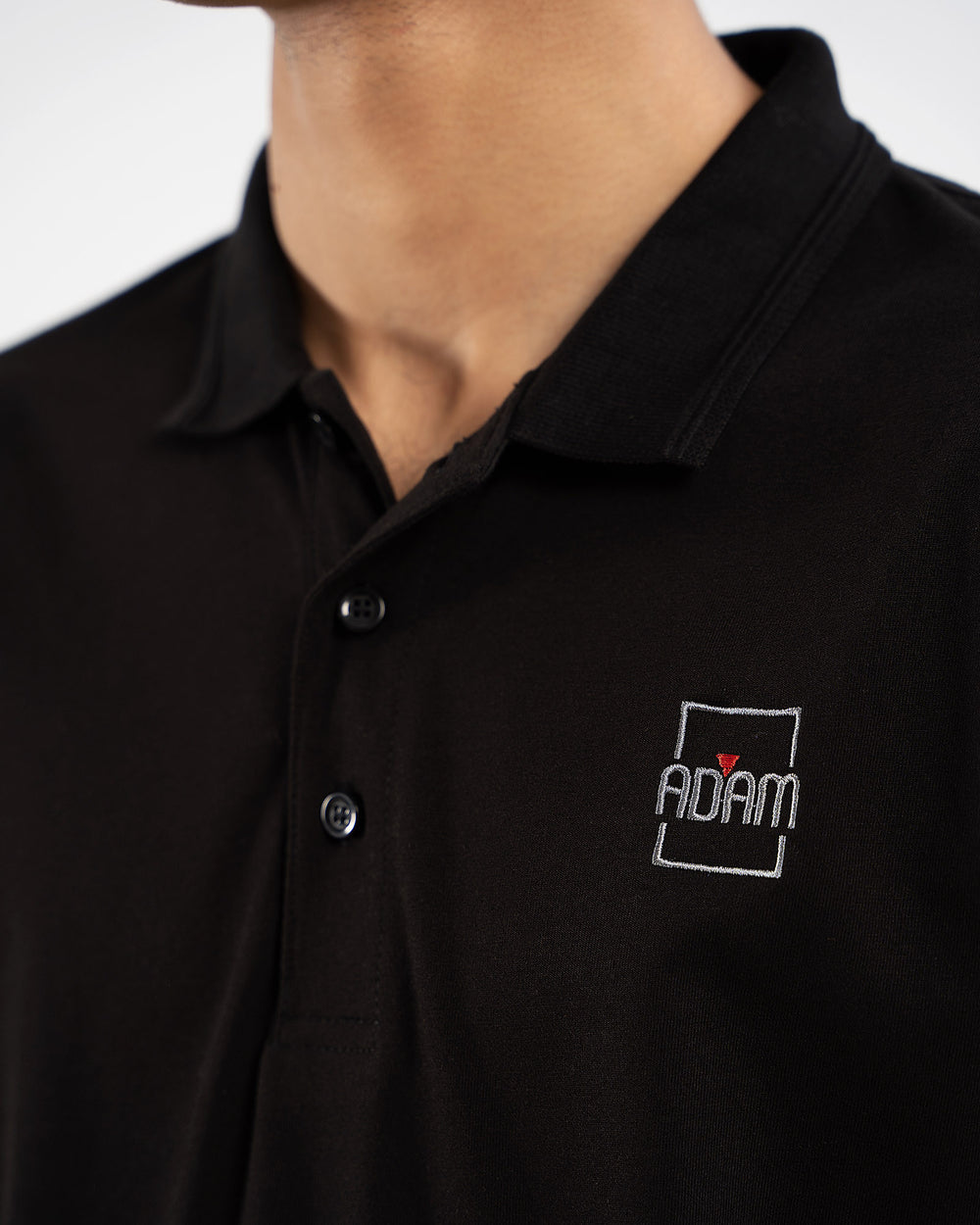 Close-up of black polo shirt with small embroidered "Adam" square box logo patch on chest