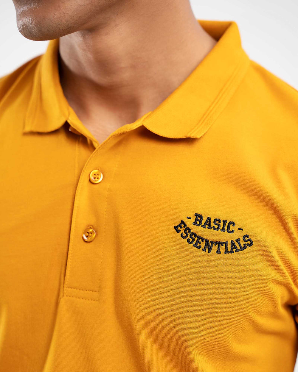 Close-up of mustard yellow polo shirt collar with small dark embroidered "Basic Essentials" arched text logo on chest
